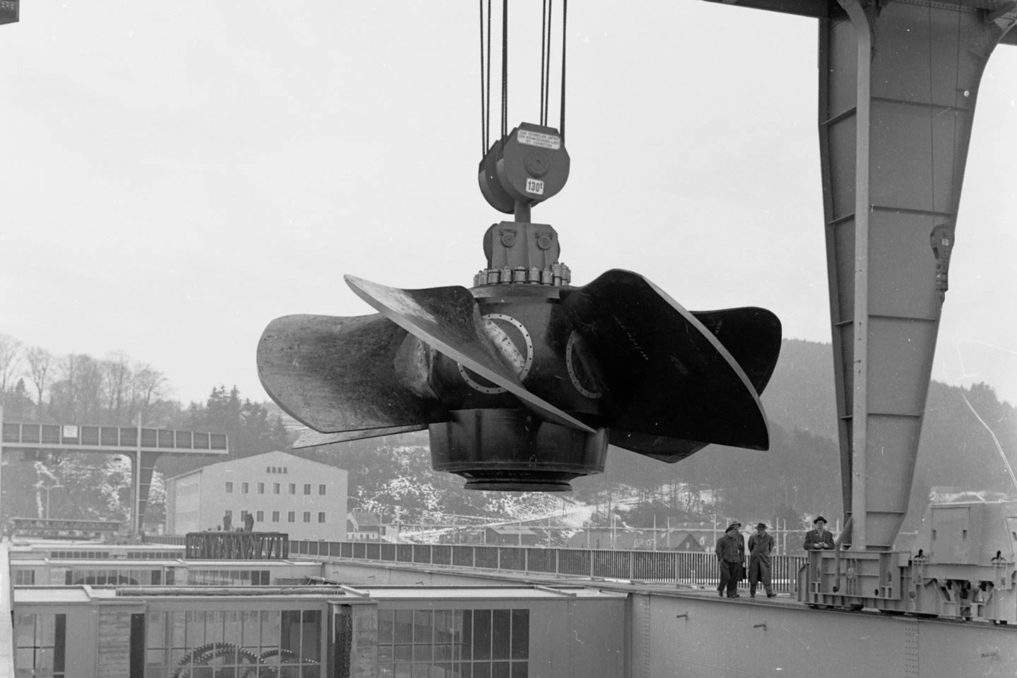 You can see a construction site photo from the past in black and white. A crane is lifting the turbine into the power station. Three men in coats and hats are walking on the pedestrian crossing of the dam wall.