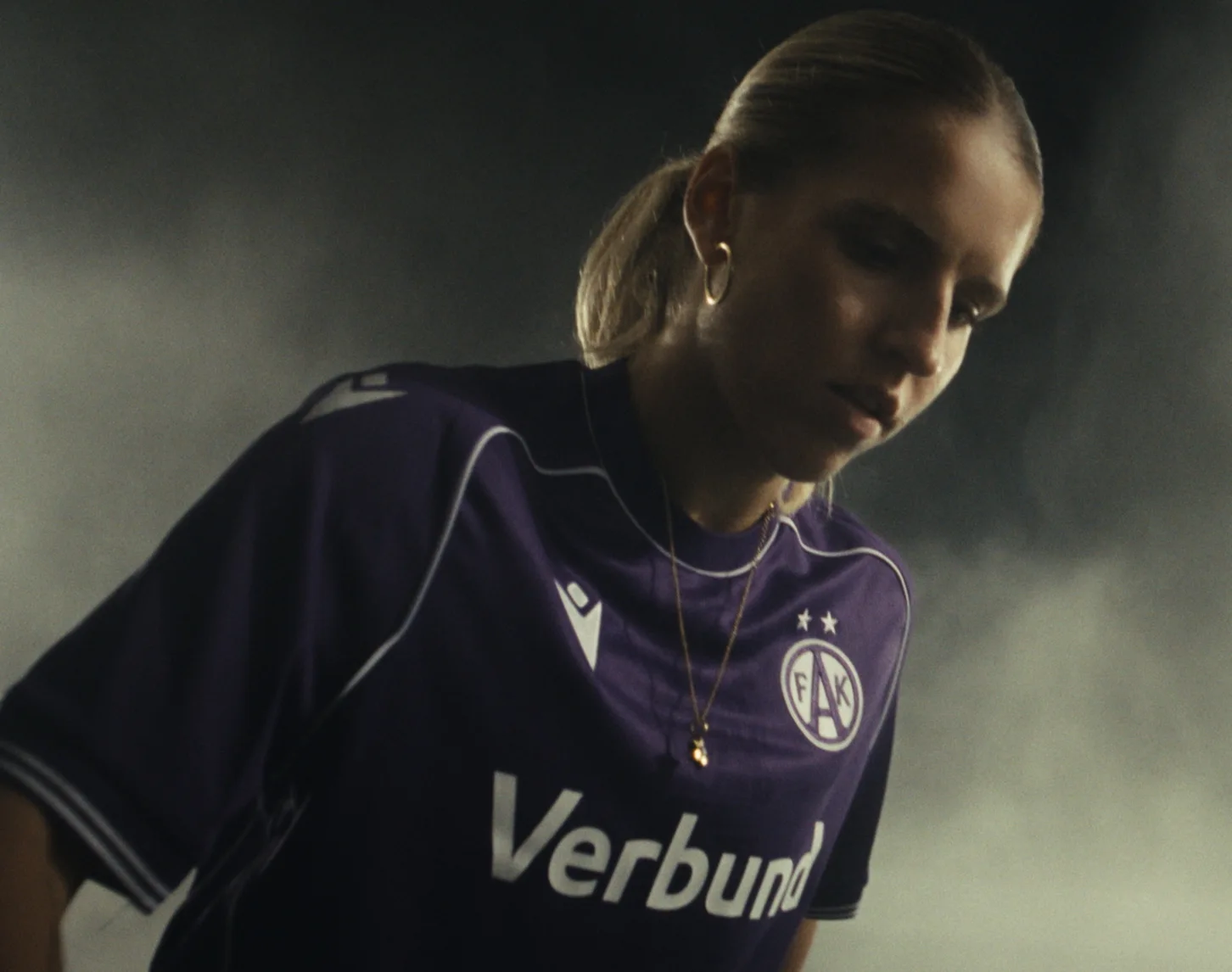 Eine Fußballspielerin im violetten FAK-Trikot mit dem Schriftzug „Verbund“ steht vor dunklem, nebligem Hintergrund und schaut nach unten.