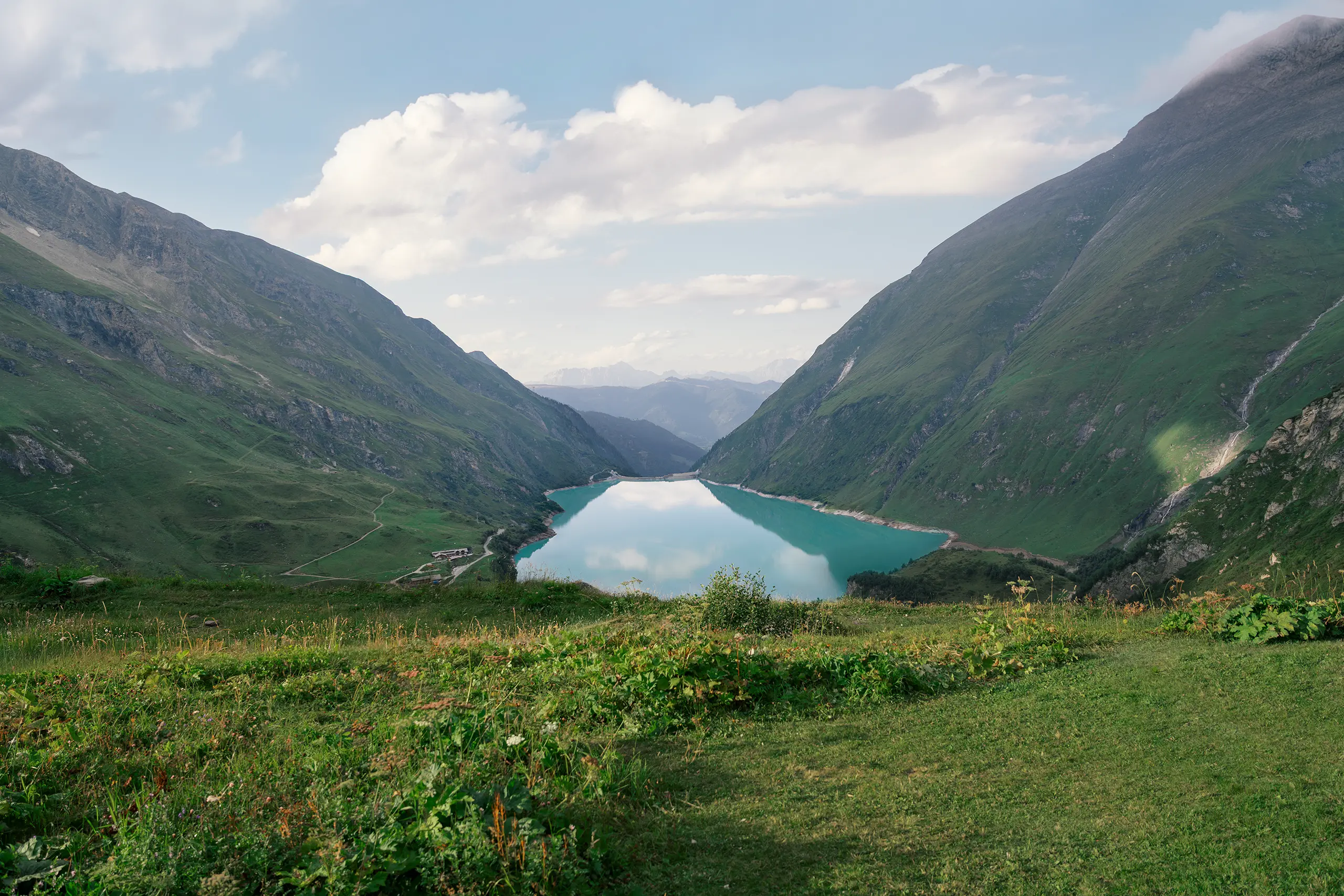 Bergtal mit türkisfarbenem Stausee zwischen grünen Hängen und Wolken am Himmel; im Vordergrund liegt eine Wiese.