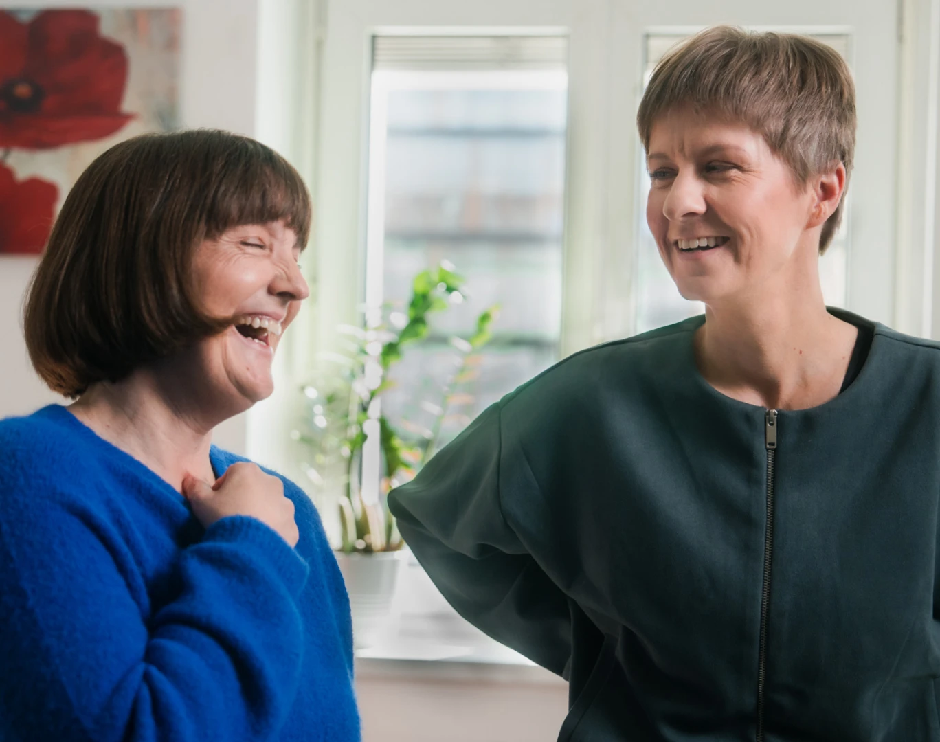 Zwei Frauen stehen zusammen und lachen. Die linke trägt einen blauen Pullover und die recht einen petrolfarbenes Oberteil.