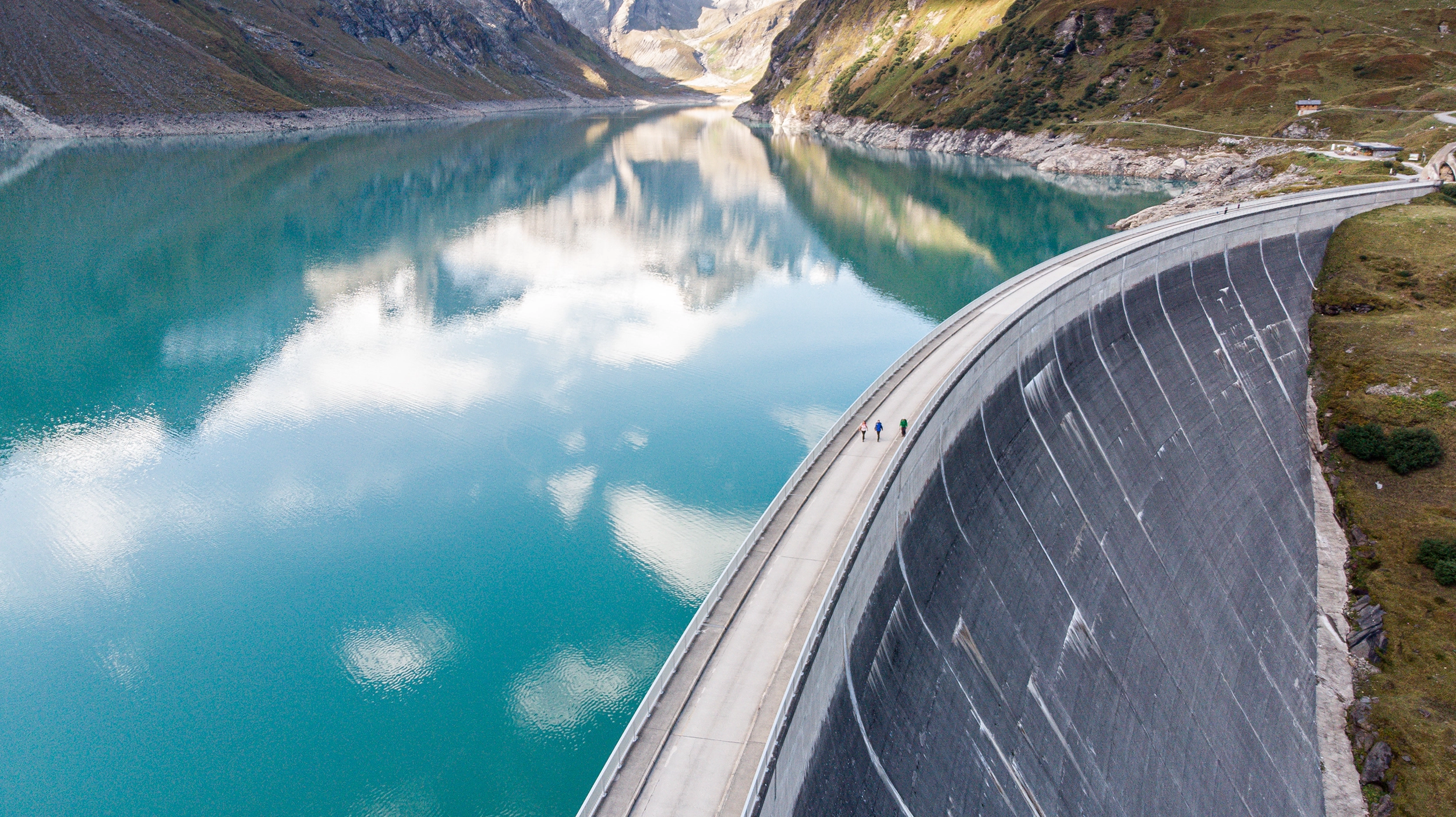 In einem herrlich blauen Stausee spiegeln sich die weißen Wolken. Am rechten Bildrand sieht man die Staumauer. Im Hintergrund spitzen die Alpen hervor.