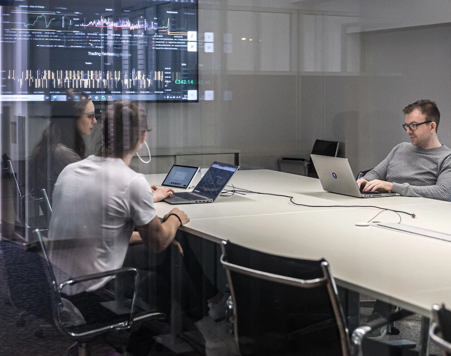 Through a transparent glass wall, you can see into a meeting room where three colleagues are sitting. Two ladies are sitting to the left of the table and a gentleman is typing something into his laptop on the right.