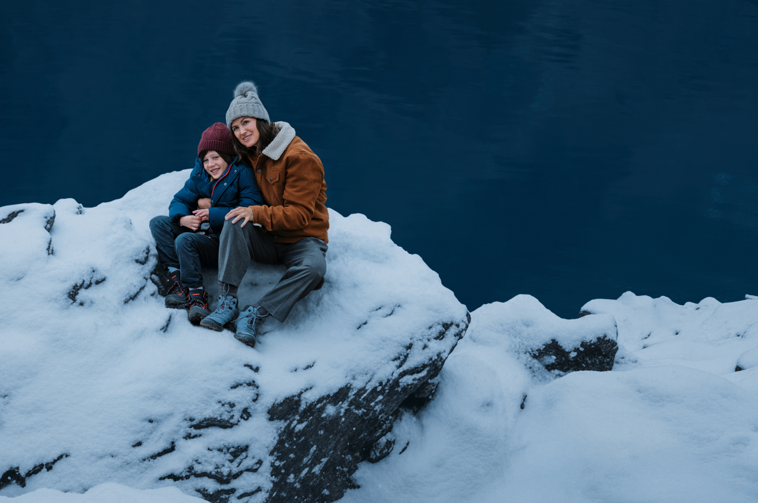 Wir blicken von oben auf Mutter uns Sohn. Beide tragen Winterklamotten und sitzen auf einem schneebedeckten Stein an einem Stausee. Sie lächeln und blicken in den Himmel.