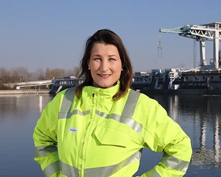 Evelin Fitzinger is standing in front of a hydroelectric power station in the sunshine, wearing a bright yellow jacket with silver reflective stripes and smiling at the camera.