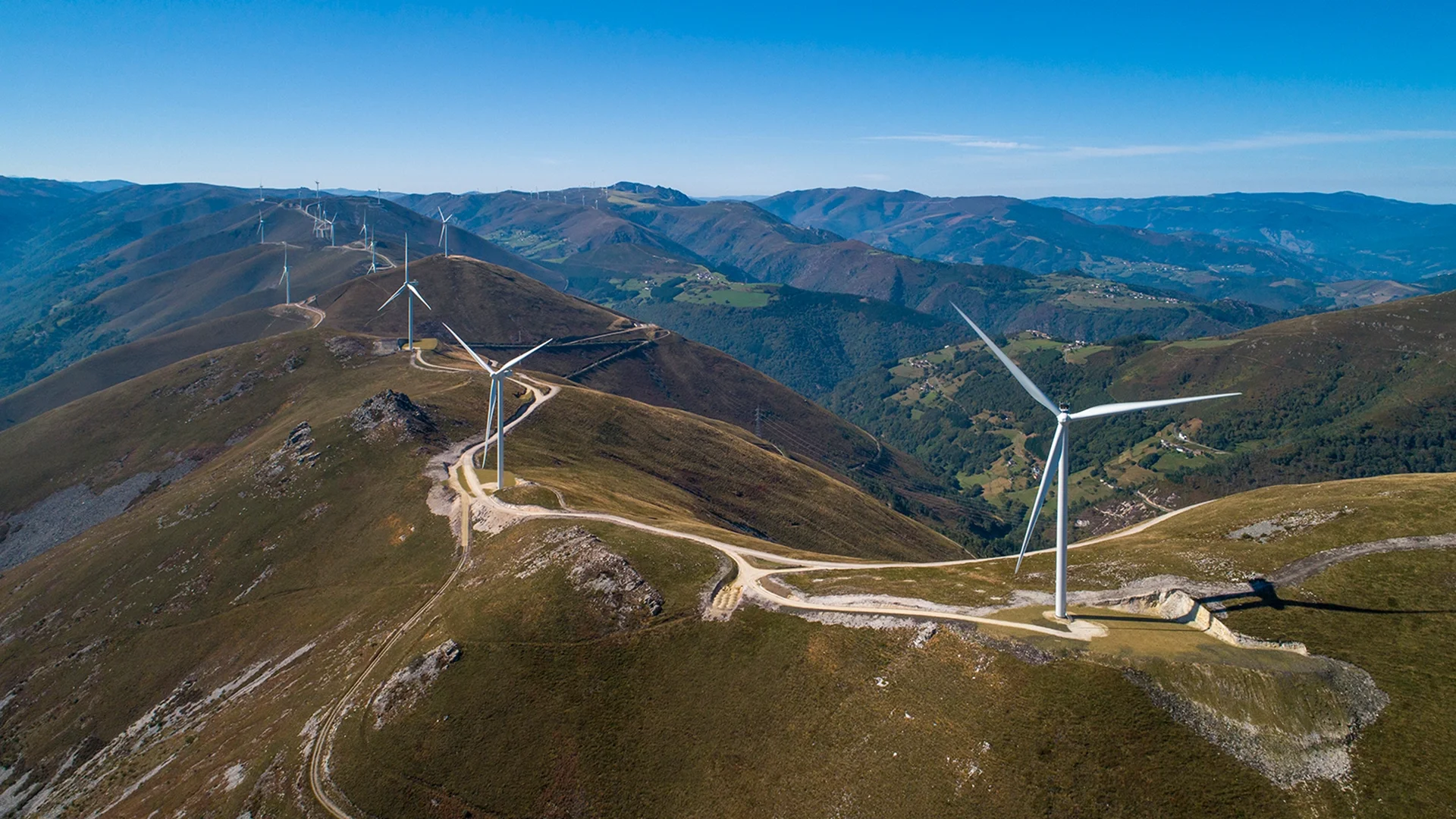 Wir blicken auf einen Windfarm in Spanien. Der Boden ist eher karg. Der Himmel ist wolkenlos und strahlende blau.