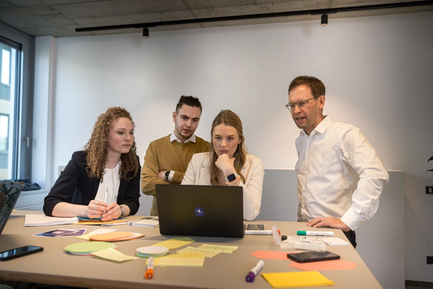 Eine Gruppe VERBUND-Mitarbeiter haben sich in einem Meetingraum versammelt, um etwas gemeinsam zu erarbeiten. Die Gruppe besteht aus zwei Männern und zwei Frauen.