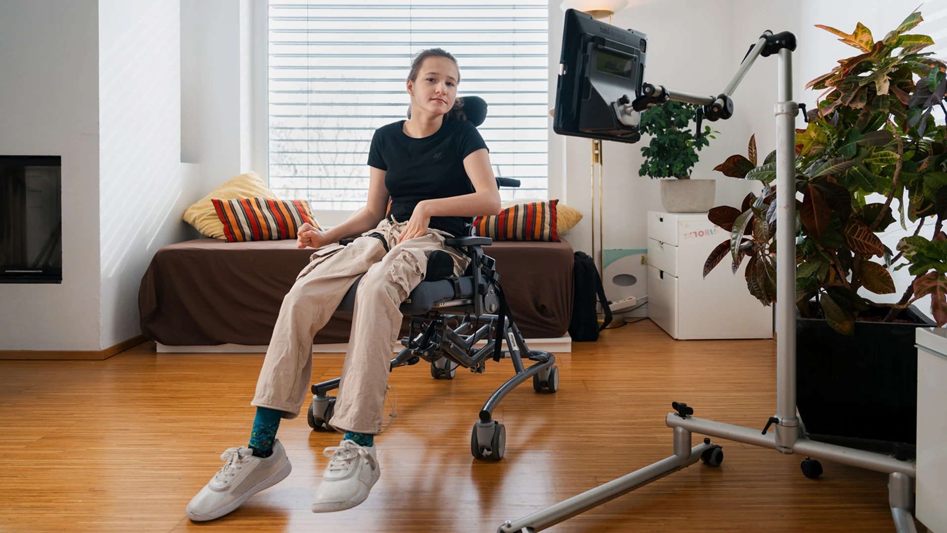 Im Mittelpunkt des Fotos sitze ein Mädchen und blick in die Kamera. Sie trägt ein dunkles T-Shirt und eine helle Hose. Am rechten Bildrand sehen sehen wir die Rückseite eines Screens.