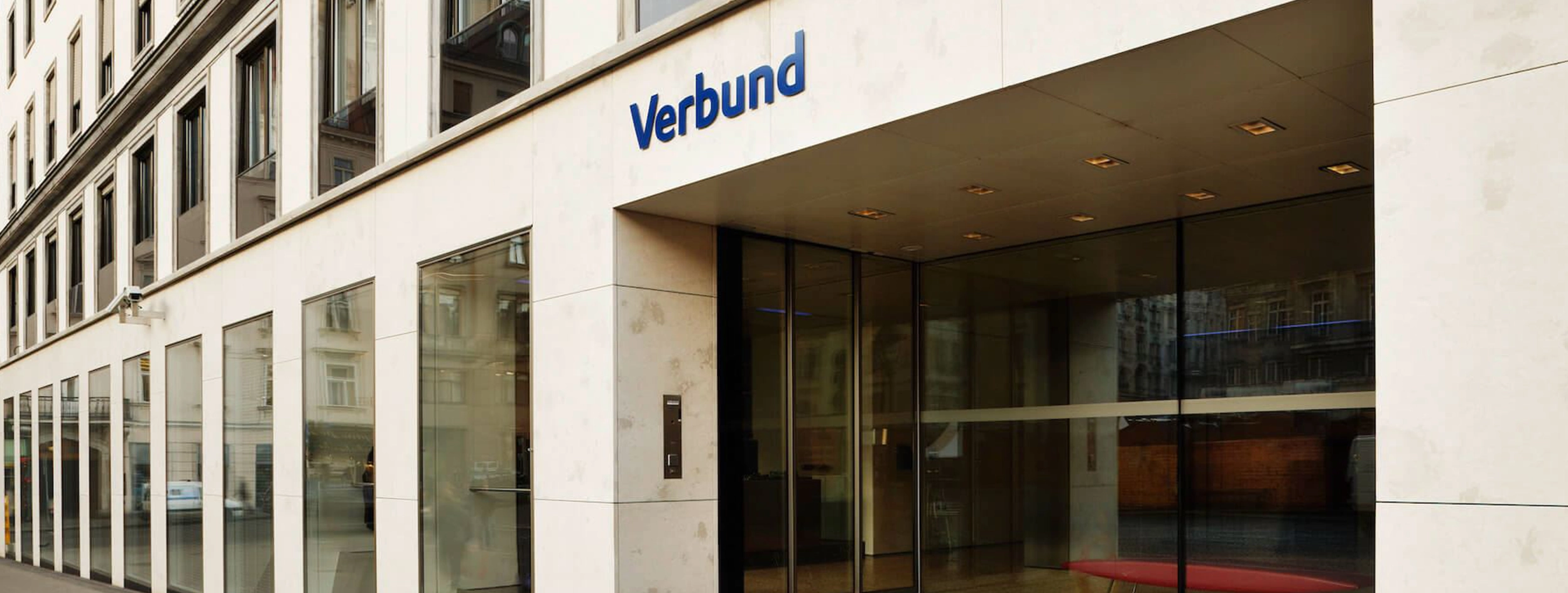 Zu sehen ist der Eingangsbereich der VERBUND Zentrale Am Hof 6A. Das Haus hat eine beige Front mit hohen Fenstern, rechts ist der Eingangsbereich mit Glasfront, darüber das VERBUND Logo.