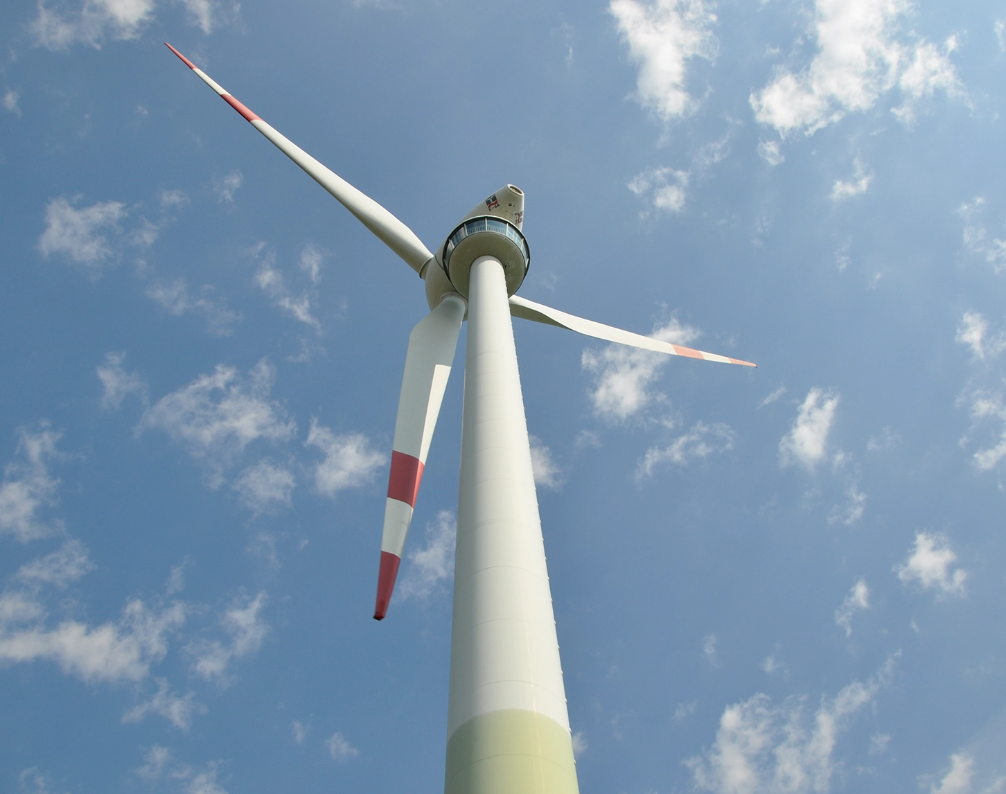We look down on the edge of the show wind in Bruck an der Leitha. The sky is blue and you can see small clouds. The wind turbine is mostly white. The tips of the wind turbines are red. Below the turbine, you can see the visitor area.