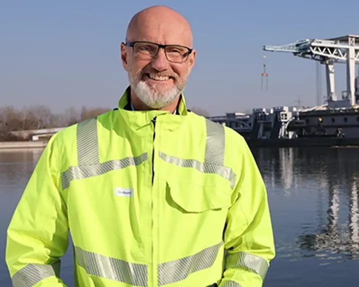 Rainer Tschopp is wearing a protective jacket in bright yellow with silver reflective stripes. He also has glasses and is standing in front of a power station in the sunshine, smiling at the camera.