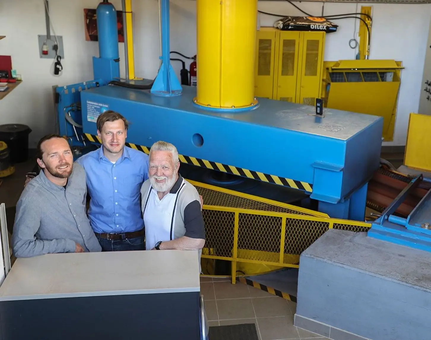 Wir sehen drei lächelnde Männer unterschiedlicher Altersgruppen. Sie stehen vor einer Art Generater oder Turbine.