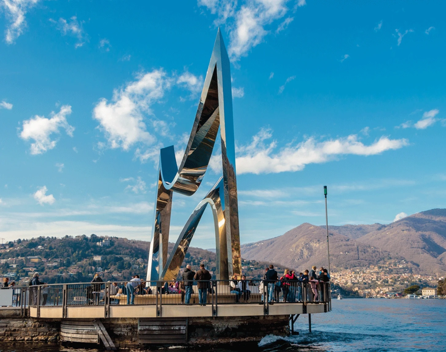 Auf dem Bild sehen wir einen steinernen Steg mit einer runden Plattform, auf der ein Monument zu ehren von Alessandro Volta steht. Es befinden sich einige Menschen auf der Plattform.