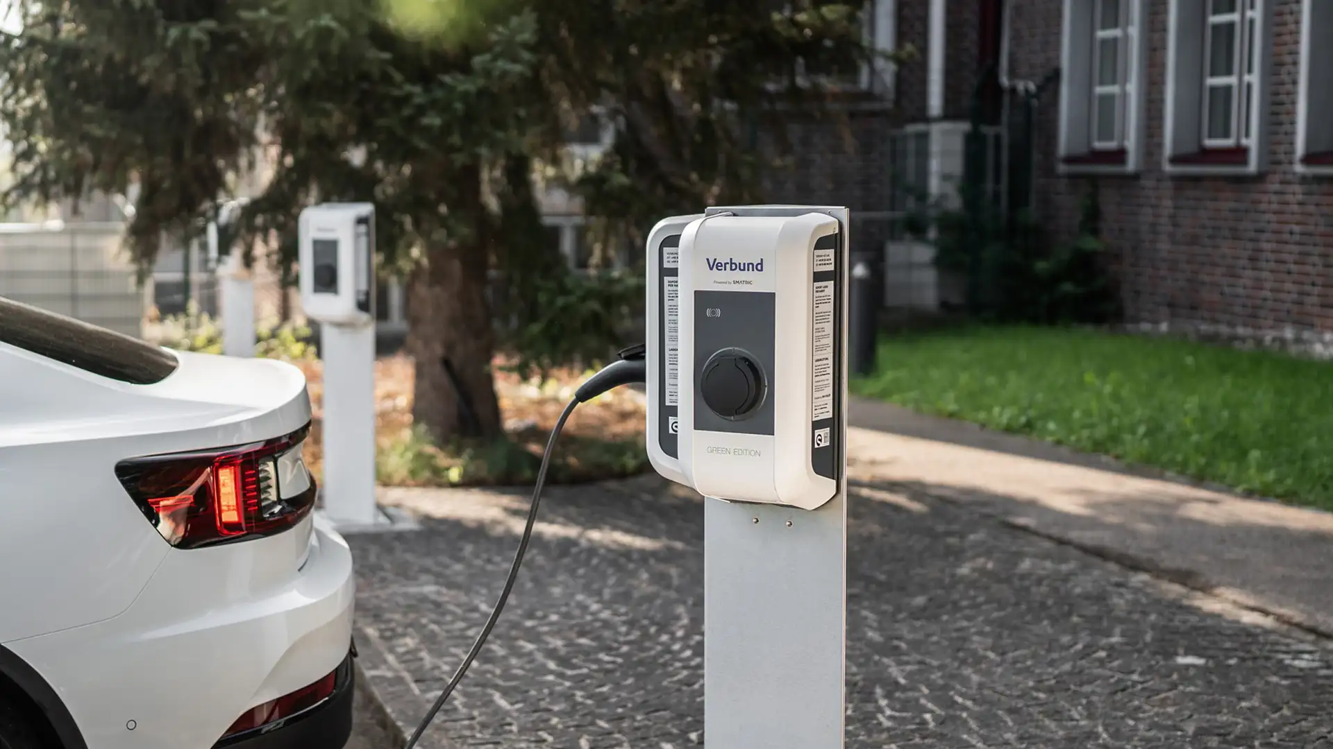 Ein E-Auto wird an einer VERBUND Ladestation auf dem Parkplatz eines Unternehmens aufgeladen, mit mehreren Ladepunkten und Bäumen in der Nähe.