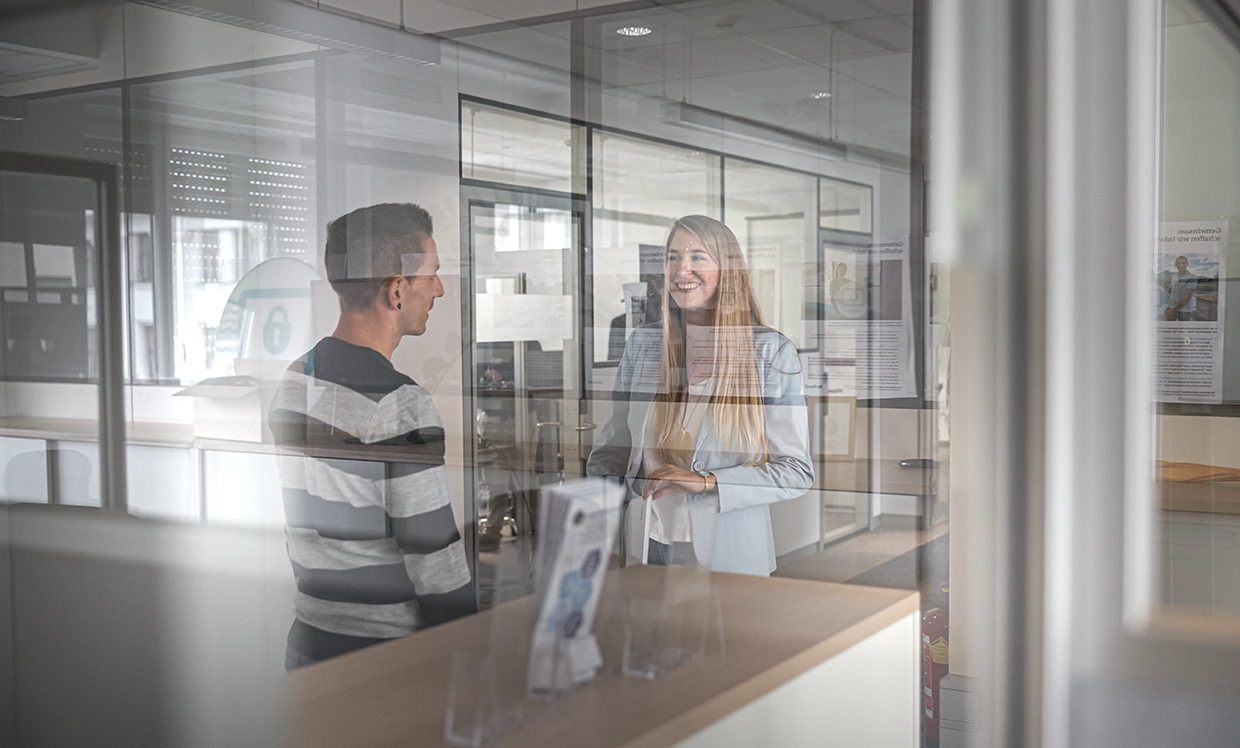 Two VERBUND employees are talking in a new office. We are separated from them by a pane of glass.