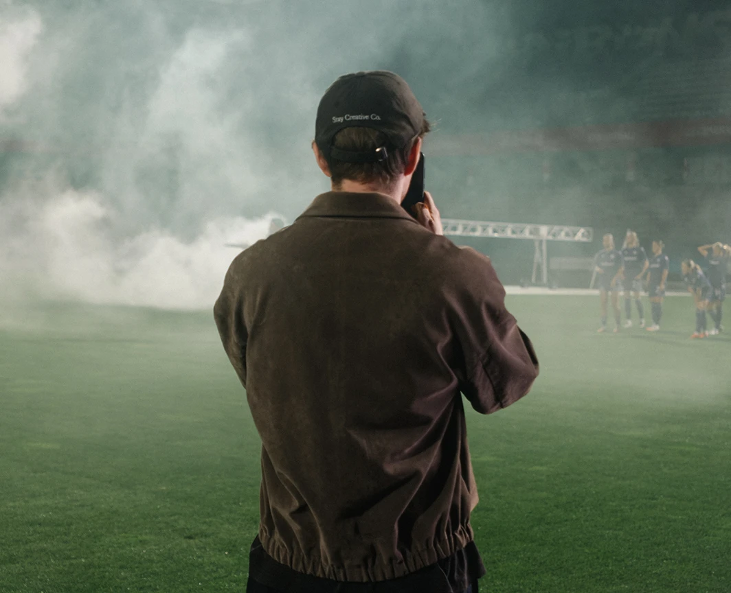 Ein Mann mit schwarzer Kappe und brauner Jacke filmt ein Team auf einem nebligen Spielfeld.