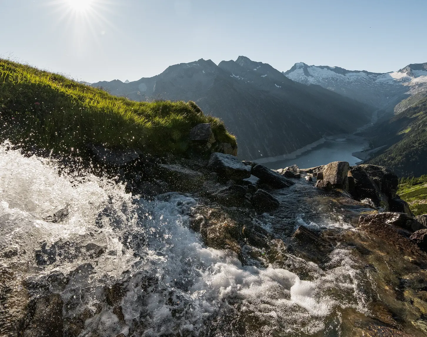 A mountain stream makes its way down into the depths. The sun is shining and the sky is blue.