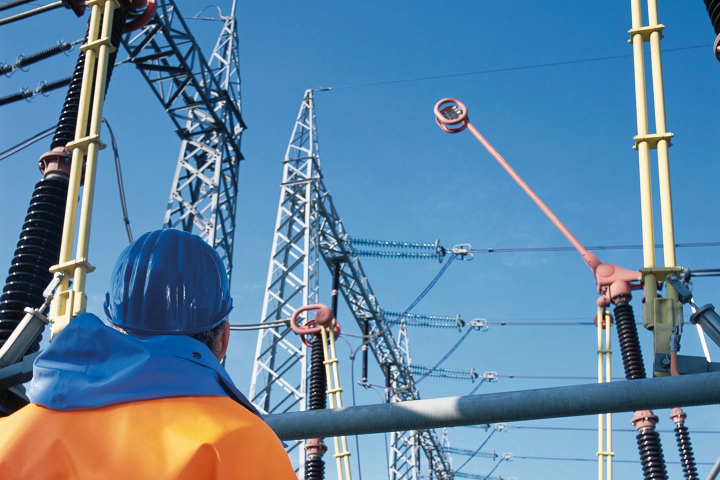 The picture shows a technical employee wearing orange protective clothing and a blue helmet. He is looking at the power lines of a transformer station. The blue sky can be seen behind him.
