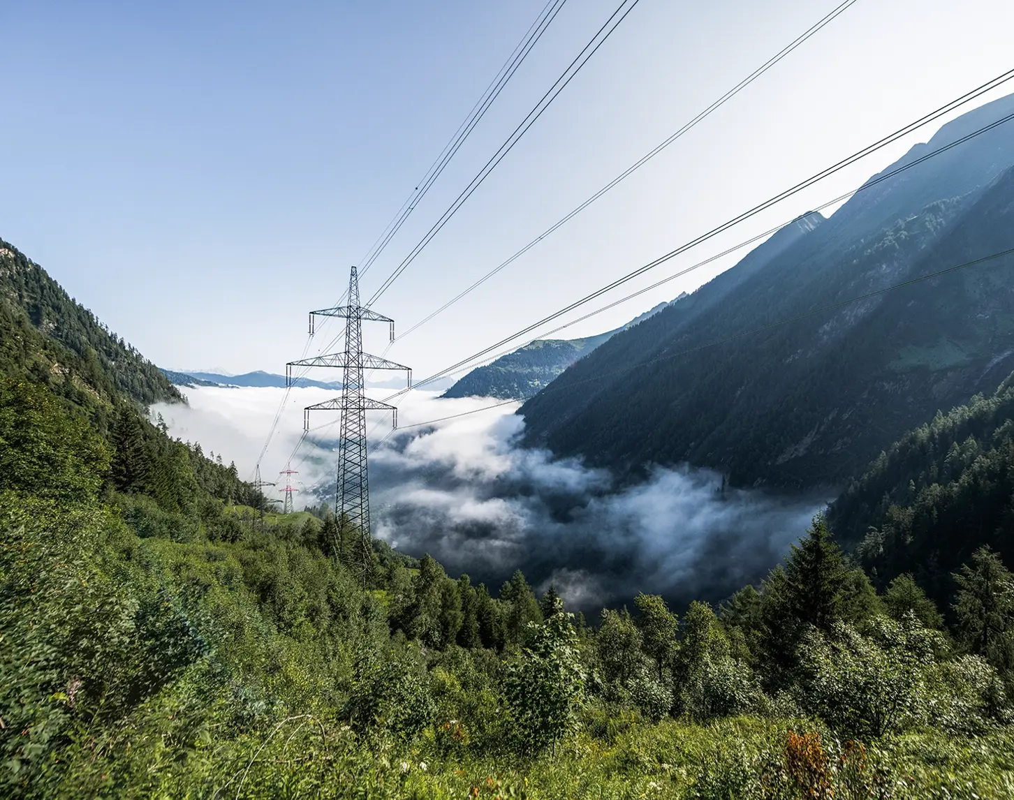Side clouds rise up between partially cloudy hills. A high-voltage grid can be seen in between.