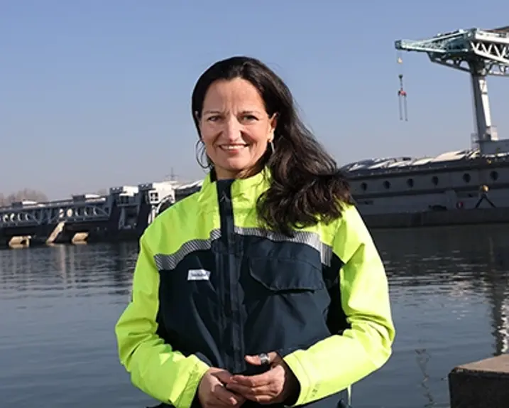 Ingun Metelko has long black hair, which she wears loose. She smiles at the camera and wears a protective jacket in yellow and dark blue with silver reflective stripes. Ingun Metelko is standing in front of a hydroelectric power station in sunny weather.