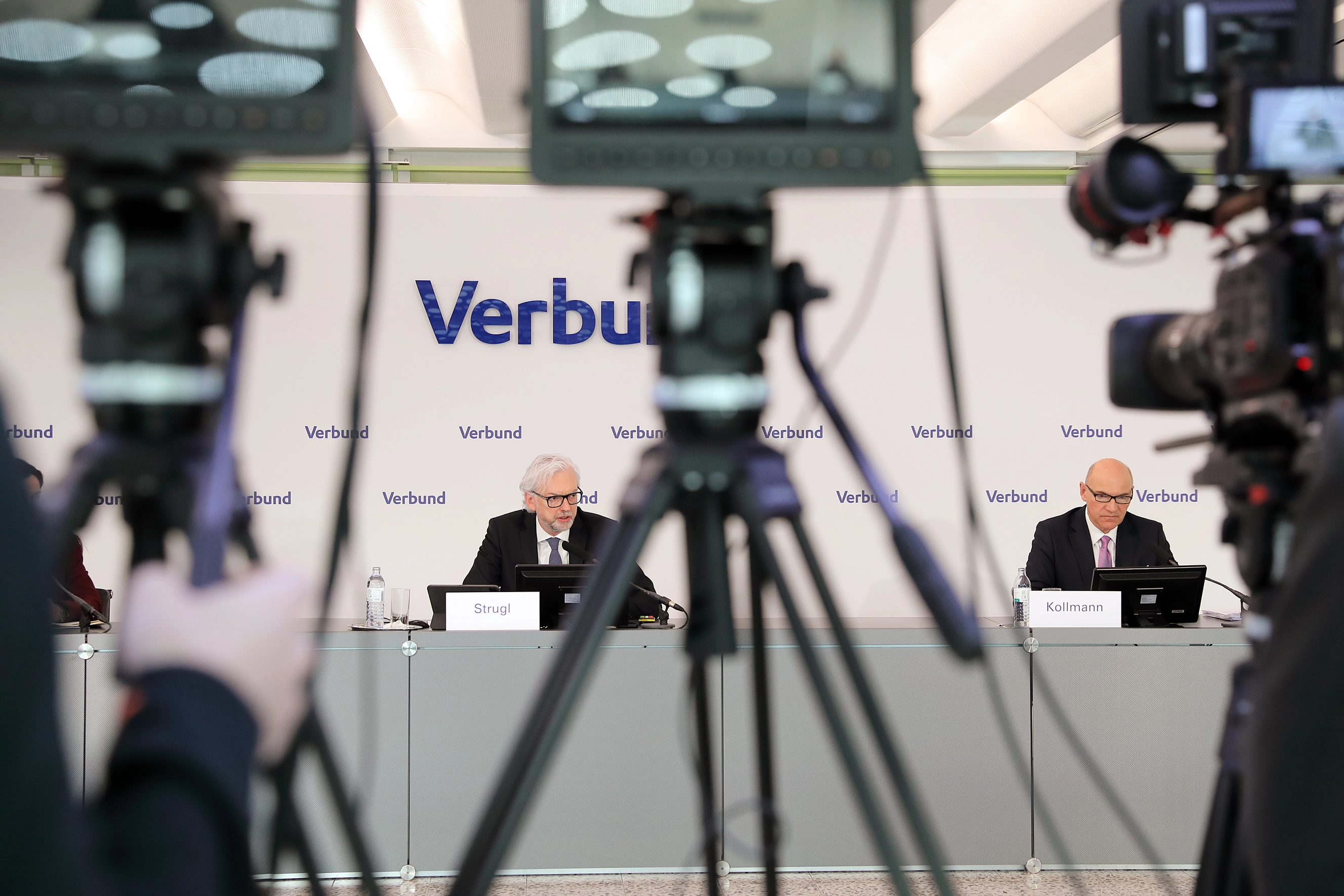 Durch die Kameras blicken wir auf Michael Stugle, CEO und Peter Kollmann, CFO von VERBUND bei einer Pressekonferenz. Wobei die Kameras in Vordergrund unscharf sind.